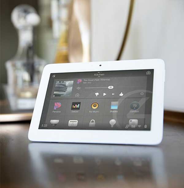 A white tablet displaying a smart home design interface, including options for music, lighting, security, and temperature, is placed on a wooden surface with a blurred background. This sleek device exemplifies the future of custom electronics and lighting integrations.
