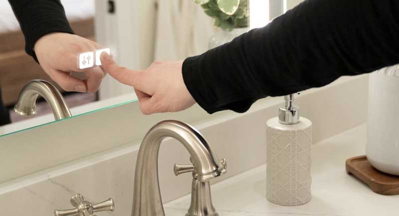 A person touching illuminated buttons on a bathroom mirror, next to a sink and soap dispenser, showcasing custom electronics and lighting integrations for a seamless smart home design.